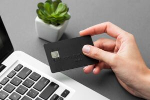 A hand holding a black credit card near a laptop keyboard with a small succulent plant in the background, symbolizing building credit in the U.S.